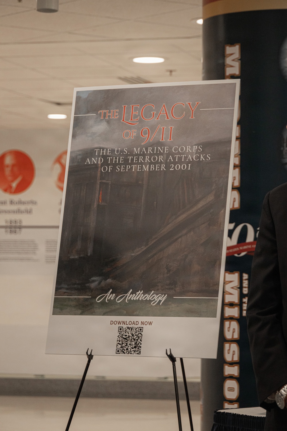 The image shows a display easel holding a promotional poster for 'The Legacy of 9/11: The U.S. Marine Corps and the Terror Attacks of September 2001 - An Anthology.' The poster features a dark, painterly background image depicting destruction and includes a QR code at the bottom labeled 'Download Now.' The setting appears to be an indoor exhibition or event space.