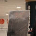 The image shows a display easel holding a promotional poster for 'The Legacy of 9/11: The U.S. Marine Corps and the Terror Attacks of September 2001 - An Anthology.' The poster features a dark, painterly background image depicting destruction and includes a QR code at the bottom labeled 'Download Now.' The setting appears to be an indoor exhibition or event space.