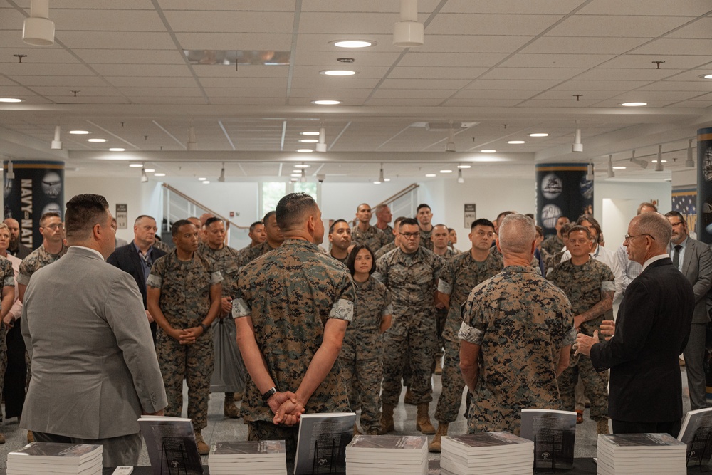 A large group of U.S. Marines in MARPAT camouflage uniforms are gathered in what appears to be a modern institutional building, listening to a civilian speaker on the right side of the frame. Stacks of books or publications are visible on tables in the foreground, suggesting a book distribution or briefing event. Two civilians in suits and one senior military figure appear to be addressing the assembled Marines.
