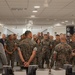 A large group of U.S. Marines in MARPAT camouflage uniforms are gathered in what appears to be a modern institutional building, listening to a civilian speaker on the right side of the frame. Stacks of books or publications are visible on tables in the foreground, suggesting a book distribution or briefing event. Two civilians in suits and one senior military figure appear to be addressing the assembled Marines.