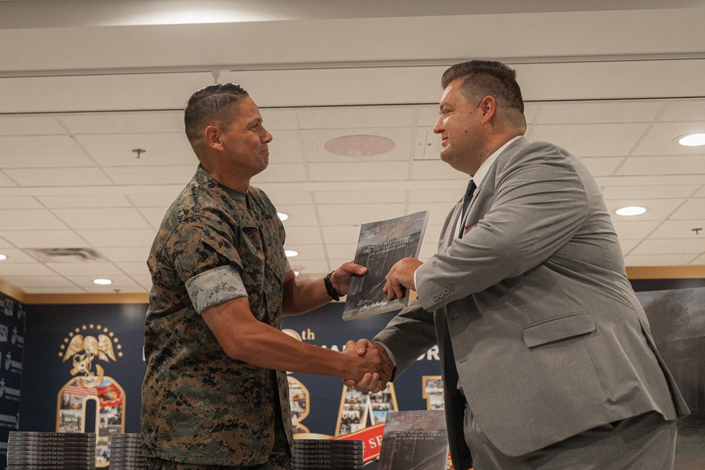 A U.S. Marine in digital camouflage uniform shakes hands with a civilian man in a gray suit during what appears to be a formal book presentation or award ceremony. The civilian is handing the Marine a publication that appears to be titled 'The U.S. Marine Corps and the Terrorist Attacks of September 2001' or similar 9/11-related content. The event appears to take place in a military facility decorated with unit insignia and banners.