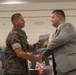 A U.S. Marine in digital camouflage uniform shakes hands with a civilian man in a gray suit during what appears to be a formal book presentation or award ceremony. The civilian is handing the Marine a publication that appears to be titled 'The U.S. Marine Corps and the Terrorist Attacks of September 2001' or similar 9/11-related content. The event appears to take place in a military facility decorated with unit insignia and banners.