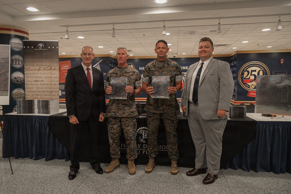 Four individuals pose for a formal photo at what appears to be a United States Marine Corps event, likely a book or publication presentation. Two individuals in the center are wearing Marine Corps MARPAT camouflage uniforms and holding what appear to be framed photographs or publications, flanked by two men in civilian business attire. The backdrop features USMC branding including a '250th Anniversary' logo and historical displays.
