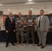 Four individuals pose for a formal photo at what appears to be a United States Marine Corps event, likely a book or publication presentation. Two individuals in the center are wearing Marine Corps MARPAT camouflage uniforms and holding what appear to be framed photographs or publications, flanked by two men in civilian business attire. The backdrop features USMC branding including a '250th Anniversary' logo and historical displays.