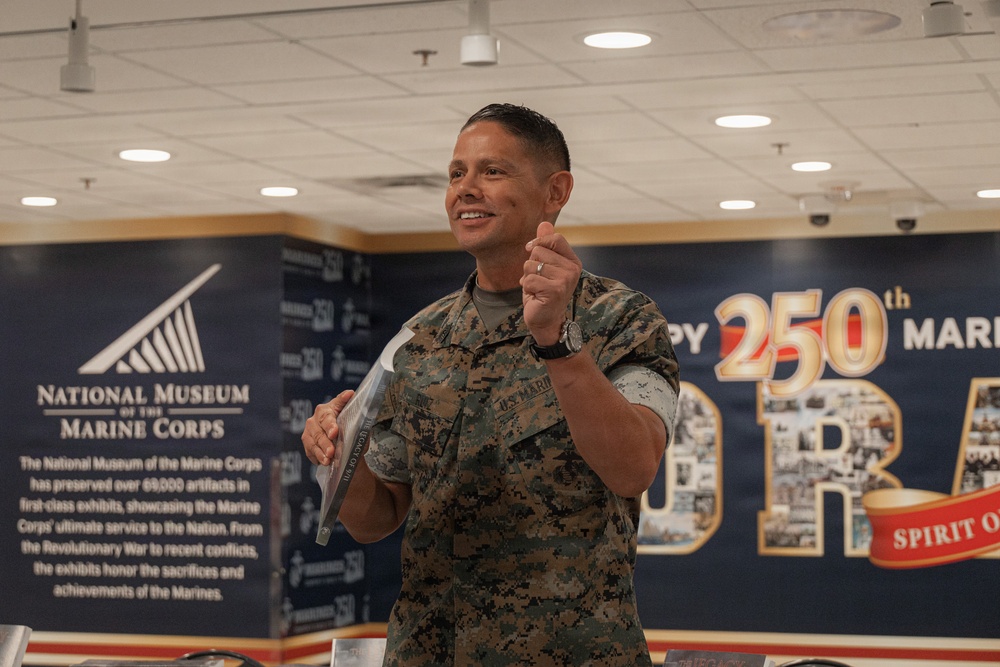 A U.S. Marine in digital camouflage uniform is speaking or presenting at an event inside what appears to be a visitor or conference center. The backdrop features signage for the National Museum of the Marine Corps and a '250th' anniversary celebration banner. The Marine appears engaged and animated, gesturing with one hand while holding documents in the other.