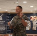 A U.S. Marine in digital camouflage uniform is speaking or presenting at an event inside what appears to be a visitor or conference center. The backdrop features signage for the National Museum of the Marine Corps and a '250th' anniversary celebration banner. The Marine appears engaged and animated, gesturing with one hand while holding documents in the other.