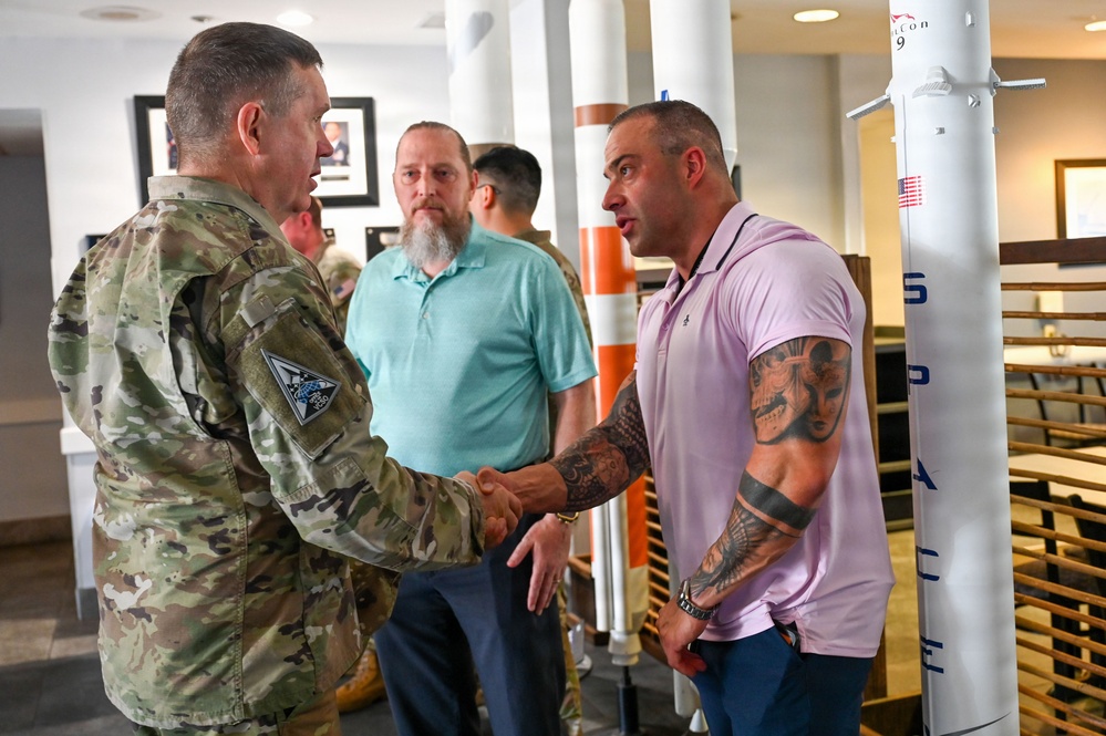 A military officer in camouflage uniform is shaking hands with a heavily tattooed civilian man wearing a pink polo shirt in what appears to be an indoor facility. A third man in a teal polo shirt observes the interaction in the background. The setting appears to be a formal or semi-formal meeting environment.