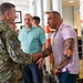 A military officer in camouflage uniform is shaking hands with a heavily tattooed civilian man wearing a pink polo shirt in what appears to be an indoor facility. A third man in a teal polo shirt observes the interaction in the background. The setting appears to be a formal or semi-formal meeting environment.