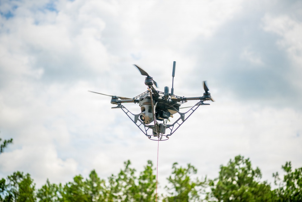 A multi-rotor drone (quadcopter or hexacopter) is captured in flight from a low-angle perspective against a partly cloudy sky. The drone is equipped with a camera gimbal payload and has a red tether line extending downward, suggesting it may be a tethered UAV system. Green treetops are visible at the bottom of the frame.