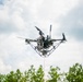 A multi-rotor drone (quadcopter or hexacopter) is captured in flight from a low-angle perspective against a partly cloudy sky. The drone is equipped with a camera gimbal payload and has a red tether line extending downward, suggesting it may be a tethered UAV system. Green treetops are visible at the bottom of the frame.