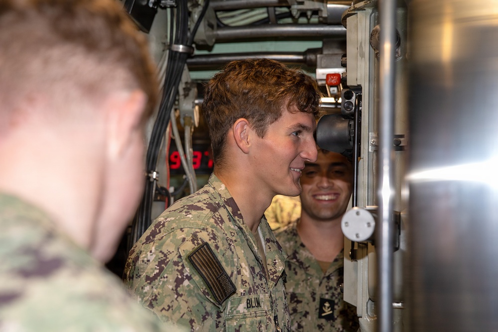 Two U.S. Navy sailors in NWU Type III (woodland digital camouflage) uniforms are visible inside what appears to be a confined naval vessel compartment, likely a submarine or ship interior. The central figure, whose name tape reads 'BLUM', is smiling and appears to be observing or operating equipment. The setting is characterized by dense machinery, cables, and metal components typical of a naval vessel interior.