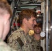 Two U.S. Navy sailors in NWU Type III (woodland digital camouflage) uniforms are visible inside what appears to be a confined naval vessel compartment, likely a submarine or ship interior. The central figure, whose name tape reads 'BLUM', is smiling and appears to be observing or operating equipment. The setting is characterized by dense machinery, cables, and metal components typical of a naval vessel interior.