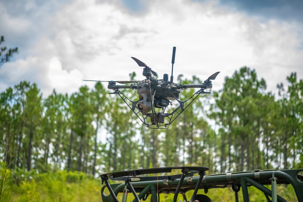 A multi-rotor drone (hexacopter or octocopter) is captured mid-flight just above what appears to be a military-green launch/landing platform. The drone is equipped with a camera gimbal system and an action camera mounted on its frame. The setting is an outdoor clearing surrounded by tall pine trees under a partly cloudy sky.