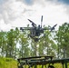 A multi-rotor drone (hexacopter or octocopter) is captured mid-flight just above what appears to be a military-green launch/landing platform. The drone is equipped with a camera gimbal system and an action camera mounted on its frame. The setting is an outdoor clearing surrounded by tall pine trees under a partly cloudy sky.