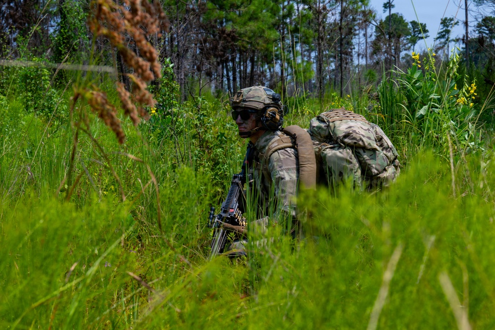 A soldier in full combat gear moves through dense, tall grass in a wooded training or operational environment. The individual is equipped with a large rucksack, tactical helmet, hearing protection, sunglasses, and carries what appears to be a belt-fed machine gun. The lush green vegetation and pine trees in the background suggest a southeastern United States environment, likely during summer.