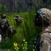 Three military personnel in full combat gear are conducting what appears to be a patrol or field training exercise along a dirt trail at the edge of a wooded area. The lead soldier is giving a hand signal, likely indicating a halt or directing movement, while the others maintain tactical spacing. The setting appears to be a subtropical or southeastern U.S. environment based on the vegetation.