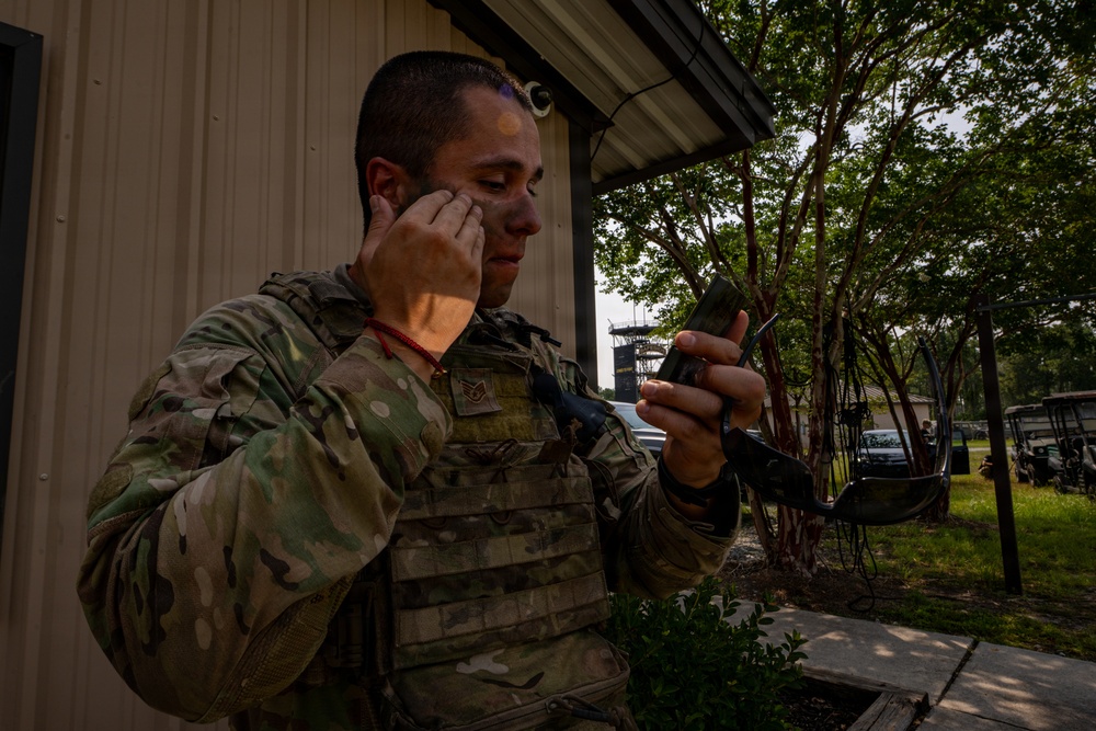 A U.S. Air Force service member in MultiCam (OCP) uniform and tactical vest is applying camouflage face paint while using a smartphone as a mirror. The soldier appears to be preparing for a field exercise or training operation, with partial face paint already applied in brown and tan tones. The setting appears to be a military training facility with trees and structures visible in the background.