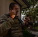 A U.S. Air Force service member in MultiCam (OCP) uniform and tactical vest is applying camouflage face paint while using a smartphone as a mirror. The soldier appears to be preparing for a field exercise or training operation, with partial face paint already applied in brown and tan tones. The setting appears to be a military training facility with trees and structures visible in the background.