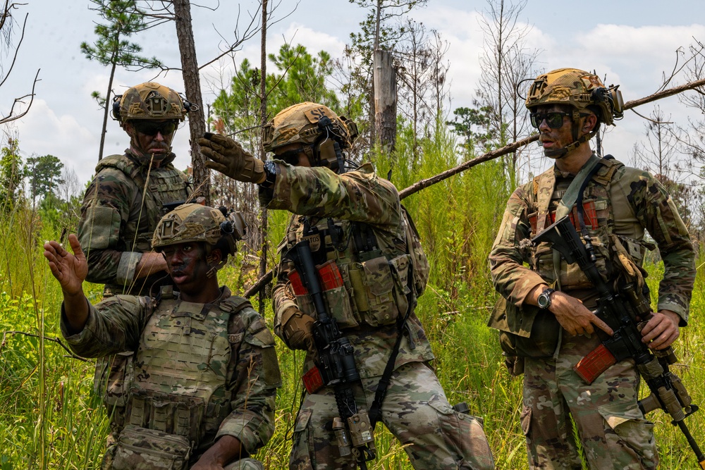 Four military personnel in full combat gear are gathered in a wooded, overgrown field environment, appearing to coordinate or brief during a field exercise or operation. They are wearing multicam/OCP camouflage uniforms with tactical vests, helmets, and communication headsets. One soldier is gesturing with his hand while another points toward a direction, suggesting active communication and coordination.