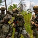 Four military personnel in full combat gear are gathered in a wooded, overgrown field environment, appearing to coordinate or brief during a field exercise or operation. They are wearing multicam/OCP camouflage uniforms with tactical vests, helmets, and communication headsets. One soldier is gesturing with his hand while another points toward a direction, suggesting active communication and coordination.