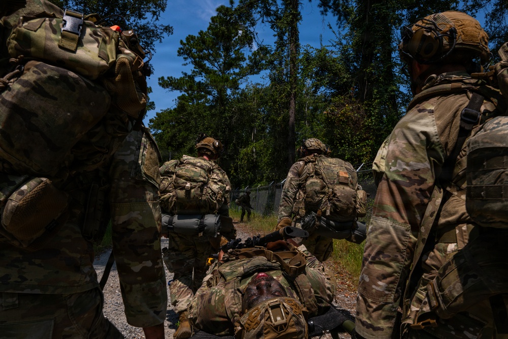 A group of military personnel in full combat gear are conducting what appears to be a tactical training exercise in a wooded outdoor environment. One soldier is lying on the ground, possibly simulating a casualty, while others stand or kneel around them in a security posture. The scene takes place along a gravel path bordered by a chain-link fence and dense tree line.