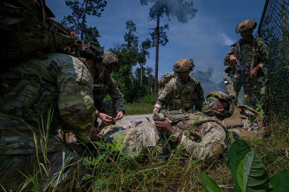 Military personnel in full combat gear are conducting a tactical combat casualty care (TCCC) exercise in a wooded outdoor environment. One soldier is positioned on the ground simulating a casualty while two or three others attend to them, performing what appears to be medical aid procedures. A fourth armed soldier stands guard in the background, maintaining security during the scenario.