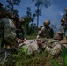 Military personnel in full combat gear are conducting a tactical combat casualty care (TCCC) exercise in a wooded outdoor environment. One soldier is positioned on the ground simulating a casualty while two or three others attend to them, performing what appears to be medical aid procedures. A fourth armed soldier stands guard in the background, maintaining security during the scenario.