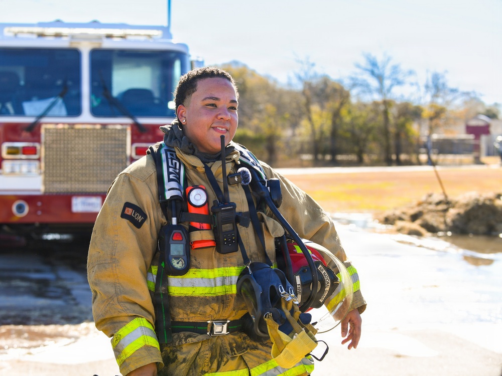 A firefighter in full turnout gear stands confidently outdoors near a red fire truck. The individual is wearing a tan/gold LION-brand bunker suit with high-visibility yellow reflective stripes and is carrying a helmet. Multiple pieces of equipment are visible on their chest, including a Motorola radio, flashlight, and other tools.