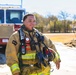 A firefighter in full turnout gear stands confidently outdoors near a red fire truck. The individual is wearing a tan/gold LION-brand bunker suit with high-visibility yellow reflective stripes and is carrying a helmet. Multiple pieces of equipment are visible on their chest, including a Motorola radio, flashlight, and other tools.