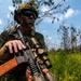 A soldier in full combat gear is positioned in a wooded, grassy area while holding an assault rifle with optical scope and accessories. The individual is wearing camouflage uniform, tactical helmet with netting, dark face paint, and reflective sunglasses. The setting appears to be a training exercise or field operation in a sparse pine forest environment.