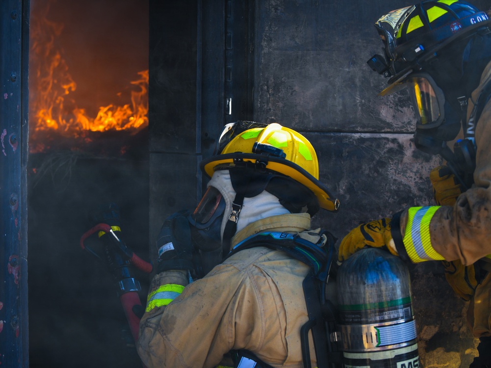 Two firefighters in full protective gear are positioned at the threshold of a burning structure, preparing to enter or manage an active fire. The scene shows intense flames visible through an opening to the left, with heavy smoke and soot covering the surrounding surfaces. The firefighters are equipped with SCBA (Self-Contained Breathing Apparatus) tanks, helmets, and turnout gear.