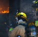 Two firefighters in full protective gear are positioned at the threshold of a burning structure, preparing to enter or manage an active fire. The scene shows intense flames visible through an opening to the left, with heavy smoke and soot covering the surrounding surfaces. The firefighters are equipped with SCBA (Self-Contained Breathing Apparatus) tanks, helmets, and turnout gear.