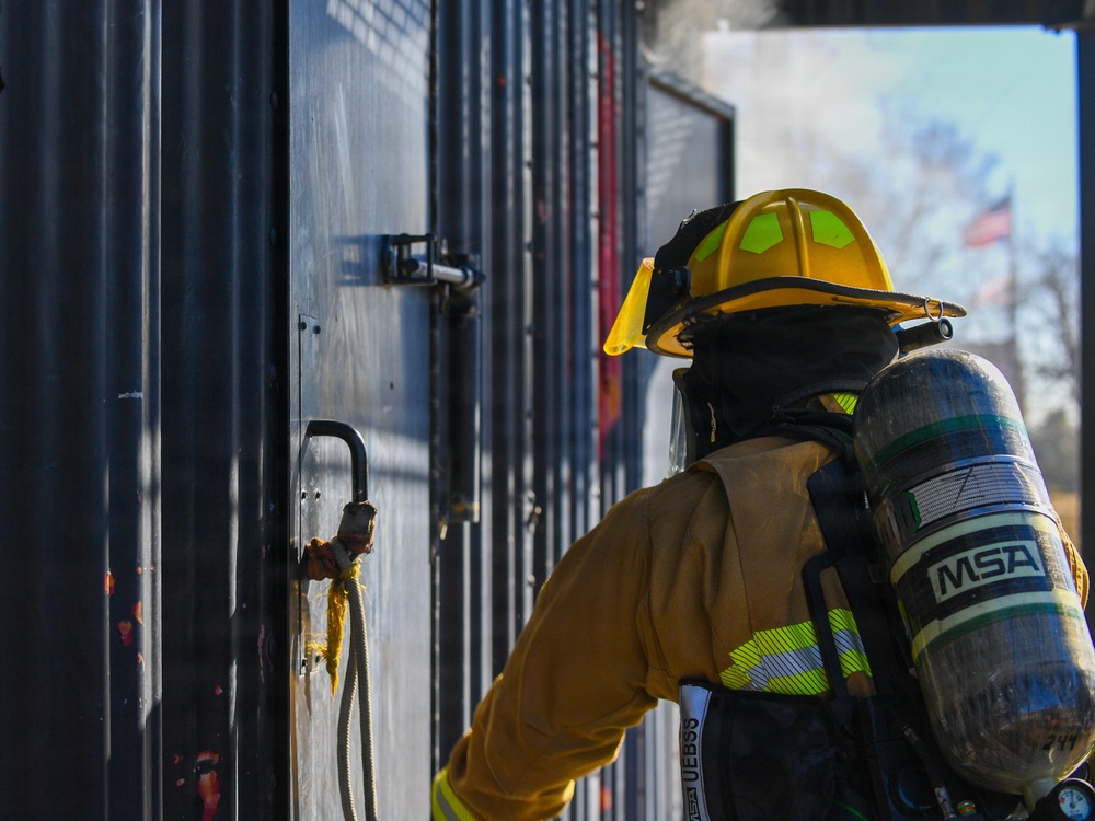 A firefighter in full turnout gear is approaching or operating a metal door, likely during a training exercise or active incident. The firefighter is equipped with a yellow helmet featuring reflective markings and an MSA self-contained breathing apparatus (SCBA) on their back. Smoke is visible in the background near an open doorway, and an American flag can be seen in the distance.