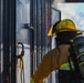 A firefighter in full turnout gear is approaching or operating a metal door, likely during a training exercise or active incident. The firefighter is equipped with a yellow helmet featuring reflective markings and an MSA self-contained breathing apparatus (SCBA) on their back. Smoke is visible in the background near an open doorway, and an American flag can be seen in the distance.