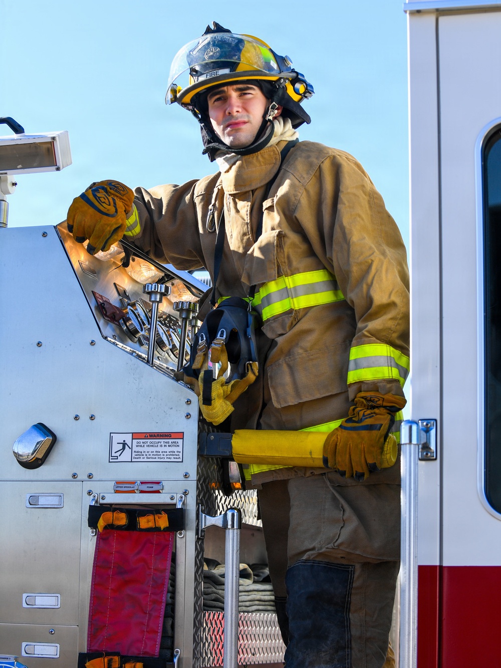 A firefighter in full turnout gear is standing on the rear step of a fire apparatus, operating or inspecting a pump panel or equipment control board. The individual is wearing a yellow helmet marked 'FIRE', tan/gold bunker gear with high-visibility lime-yellow reflective striping, and leather work gloves. The image appears to be taken during daylight under clear skies, possibly during a training exercise or pre-deployment equipment check.