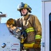 A firefighter in full turnout gear is standing on the rear step of a fire apparatus, operating or inspecting a pump panel or equipment control board. The individual is wearing a yellow helmet marked 'FIRE', tan/gold bunker gear with high-visibility lime-yellow reflective striping, and leather work gloves. The image appears to be taken during daylight under clear skies, possibly during a training exercise or pre-deployment equipment check.