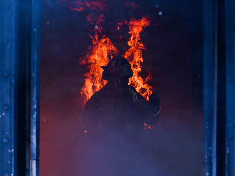 A firefighter in full protective gear and helmet stands silhouetted against large, active flames in a smoke-filled environment. The figure appears to be inside a structure or framed by a doorway or window, with intense orange and red fire burning directly behind them. The dramatic backlighting from the flames creates a stark silhouette effect against the dark, smoke-laden atmosphere.