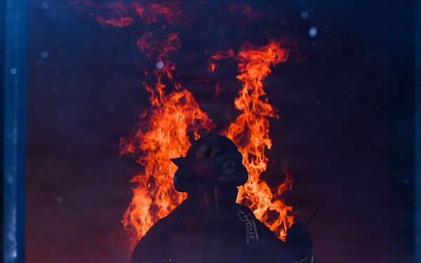 A firefighter in full protective gear and helmet stands silhouetted against large, active flames in a smoke-filled environment. The figure appears to be inside a structure or framed by a doorway or window, with intense orange and red fire burning directly behind them. The dramatic backlighting from the flames creates a stark silhouette effect against the dark, smoke-laden atmosphere.