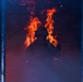 A firefighter in full protective gear and helmet stands silhouetted against large, active flames in a smoke-filled environment. The figure appears to be inside a structure or framed by a doorway or window, with intense orange and red fire burning directly behind them. The dramatic backlighting from the flames creates a stark silhouette effect against the dark, smoke-laden atmosphere.