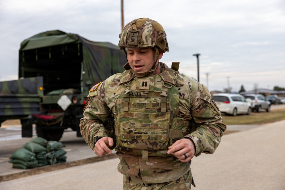 A U.S. Army soldier identified by name tape as 'ELLIS' is adjusting or securing his body armor/plate carrier in an outdoor military installation setting. He is wearing full OCP (Operational Camouflage Pattern) uniform with a tactical helmet, rank insignia visible on his chest (Captain's bars), and a U.S. flag patch on his left shoulder. Military vehicles, sandbags, and a parking area are visible in the background.
