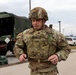 A U.S. Army soldier identified by name tape as 'ELLIS' is adjusting or securing his body armor/plate carrier in an outdoor military installation setting. He is wearing full OCP (Operational Camouflage Pattern) uniform with a tactical helmet, rank insignia visible on his chest (Captain's bars), and a U.S. flag patch on his left shoulder. Military vehicles, sandbags, and a parking area are visible in the background.