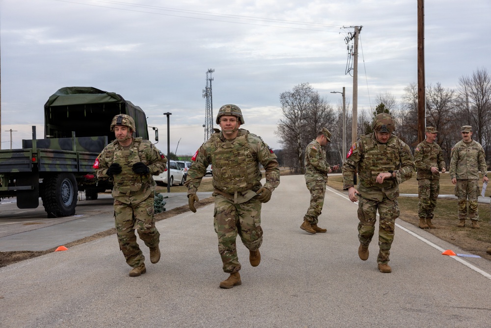 A group of military personnel in camouflage uniforms and combat gear are running or moving briskly along a paved road at what appears to be a military installation or training base. Several soldiers wear helmets and body armor with red cross or unit patches visible on their sleeves, suggesting a timed physical fitness or combat readiness evaluation. A military cargo truck with a canvas cover is parked on the left side of the road.