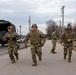 A group of military personnel in camouflage uniforms and combat gear are running or moving briskly along a paved road at what appears to be a military installation or training base. Several soldiers wear helmets and body armor with red cross or unit patches visible on their sleeves, suggesting a timed physical fitness or combat readiness evaluation. A military cargo truck with a canvas cover is parked on the left side of the road.