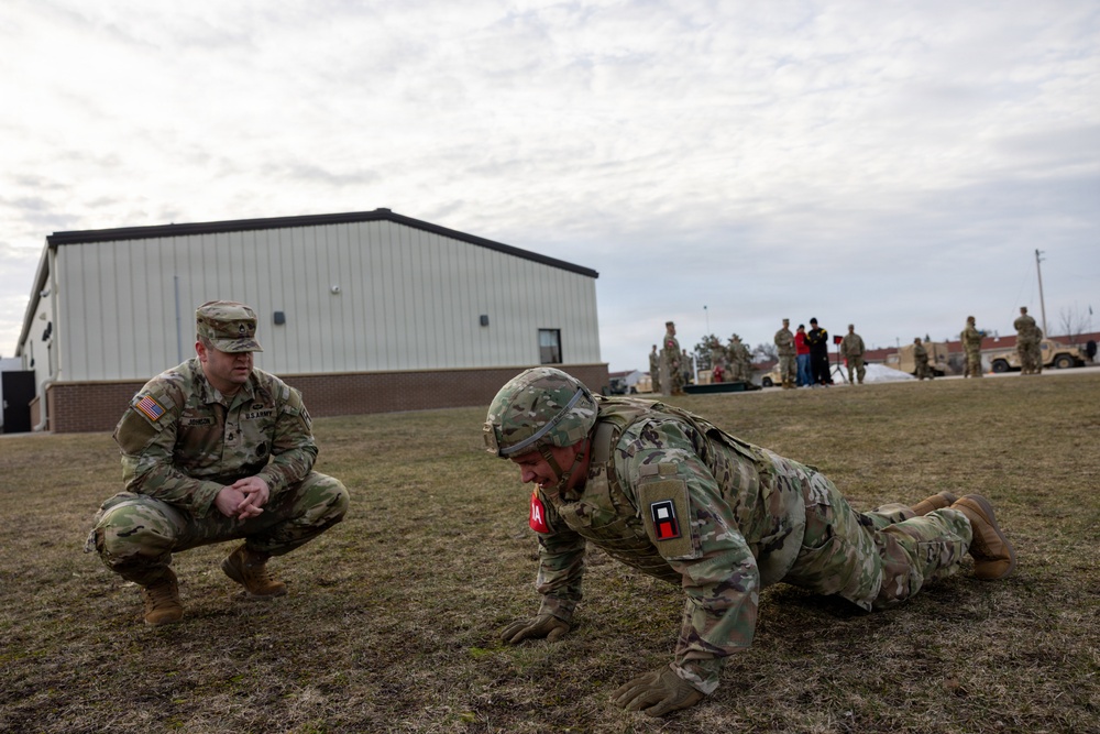 Two U.S. Army soldiers are in the foreground on a grass field, with one performing push-ups while the other squats nearby observing, likely during a physical fitness test or training exercise. Additional soldiers and military vehicles are visible in the background near a metal warehouse-style building. The setting appears to be a military installation or training area.