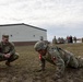 Two U.S. Army soldiers are in the foreground on a grass field, with one performing push-ups while the other squats nearby observing, likely during a physical fitness test or training exercise. Additional soldiers and military vehicles are visible in the background near a metal warehouse-style building. The setting appears to be a military installation or training area.