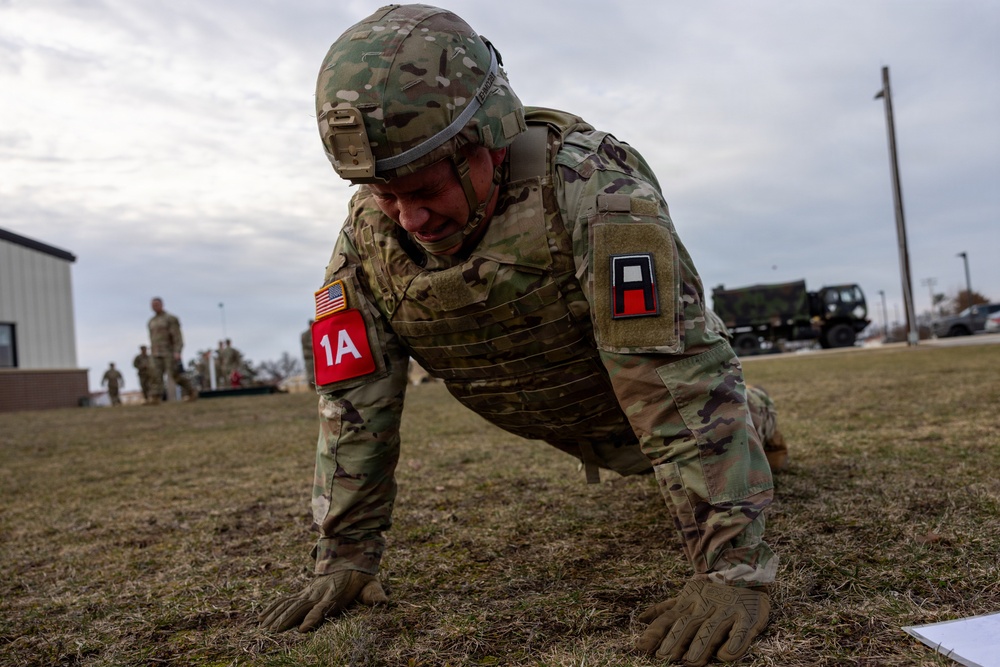 A U.S. Army soldier in full combat gear, including helmet and body armor, performs push-ups on a grass field at what appears to be a military installation. The soldier wears a '1A' patch on the left sleeve and a U.S. flag patch, indicating affiliation with First Army. Additional soldiers and military vehicles are visible in the background.
