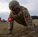 A U.S. Army soldier in full combat gear, including helmet and body armor, performs push-ups on a grass field at what appears to be a military installation. The soldier wears a '1A' patch on the left sleeve and a U.S. flag patch, indicating affiliation with First Army. Additional soldiers and military vehicles are visible in the background.