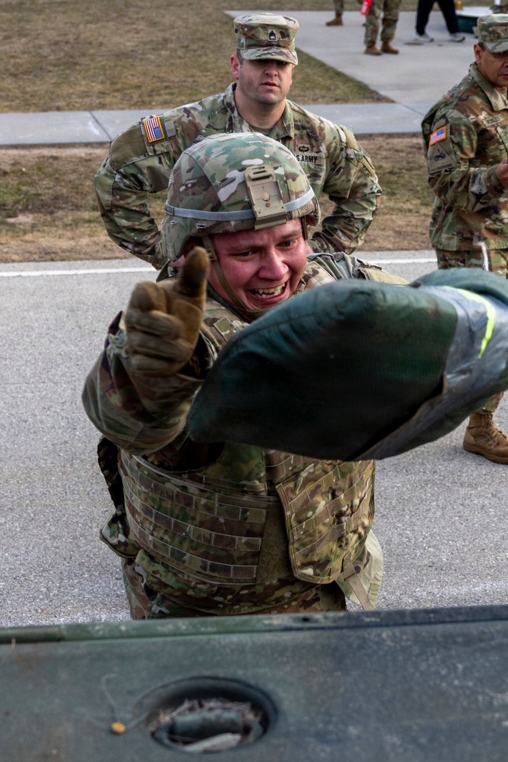 A U.S. Army soldier wearing a combat helmet and body armor is straining with visible exertion, appearing to lift or load a heavy object near a military vehicle. A non-commissioned officer (NCO) wearing a staff sergeant rank patch observes from behind. The setting appears to be a military training or equipment exercise on a paved surface.