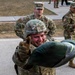 A U.S. Army soldier wearing a combat helmet and body armor is straining with visible exertion, appearing to lift or load a heavy object near a military vehicle. A non-commissioned officer (NCO) wearing a staff sergeant rank patch observes from behind. The setting appears to be a military training or equipment exercise on a paved surface.