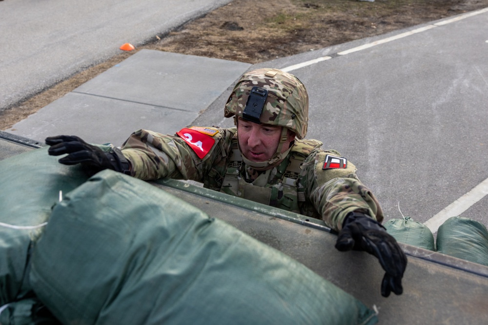 A soldier in MultiCam/OCP camouflage uniform is climbing over or peering over a barrier or vehicle edge, with green duffel bags or equipment visible in the foreground. The soldier is wearing a combat helmet with a mounted device, black tactical gloves, and shoulder patches including a red-bordered unit patch. The setting appears to be a paved military installation or training area.